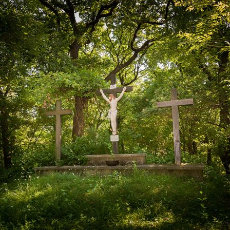 Drei Holzkreuze stehen in einer Waldlichtung. Das mittlere Kreuz hat einen Kruzifix mit Jesus Christus darauf. Das Kreuz steht auf einem Betonsockel mit einer Schale in der Nähe.