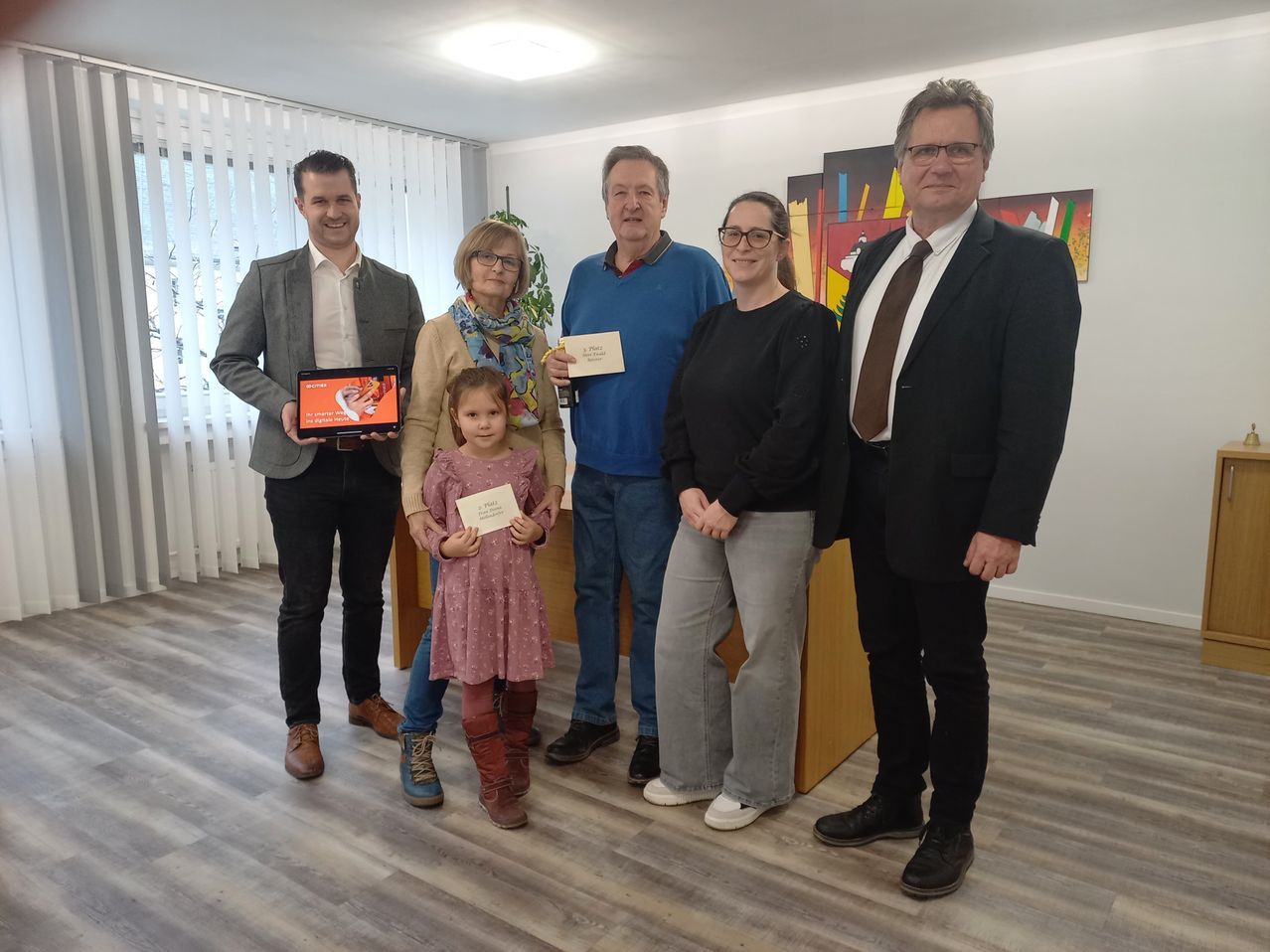 Fünf Personen stehen in einem Raum, lächeln und halten Urkunden in den Händen. Ein kleines Mädchen hält ebenfalls eine Urkunde. Sie stehen vor einem Holzschreibtisch.