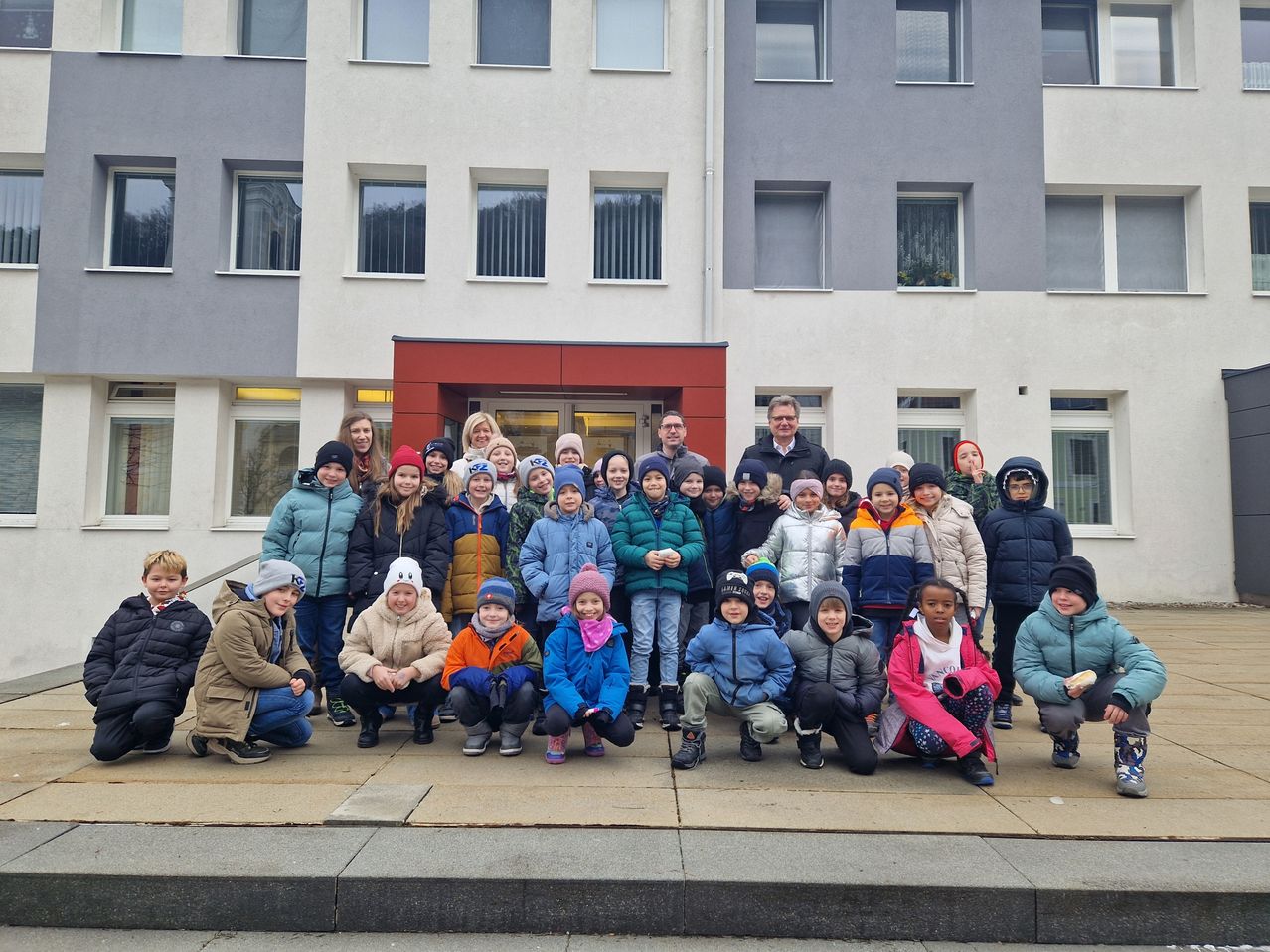 Eine Gruppe von Schülern, Lehrern und Erwachsenen in Winterkleidung posiert für ein Foto vor einem Gebäude mit mehreren Fenstern.