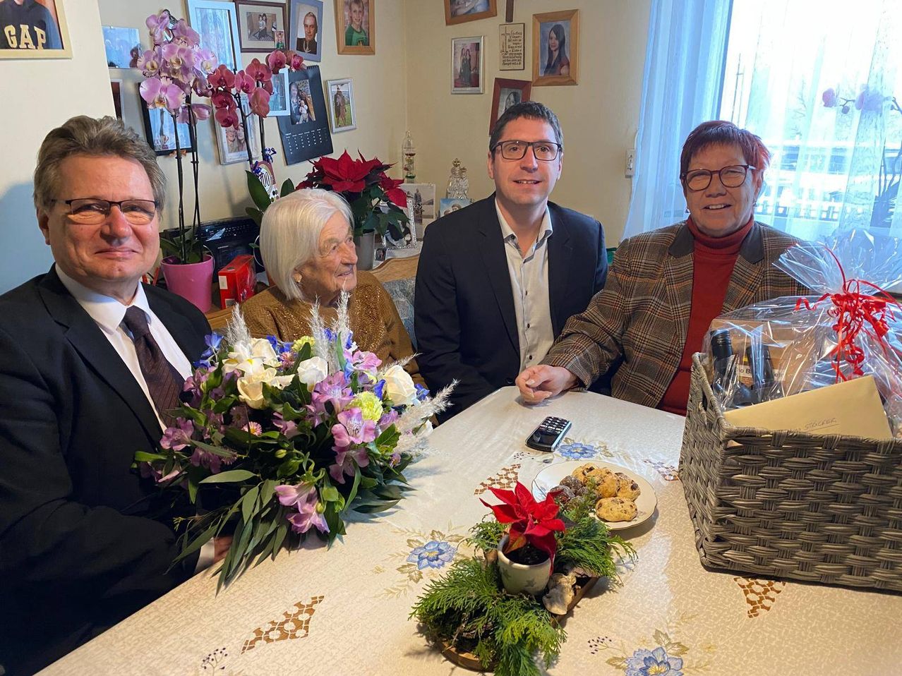 Vier Personen sitzen an einem Tisch in einem Raum mit einem Blumencenterpiece und einer Platte mit Keksen. Der Mann auf der linken Seite hält einen Blumenstrauß. Die anderen tragen formelle Kleidung. Familienfotos schmücken die Wand hinter ihnen.