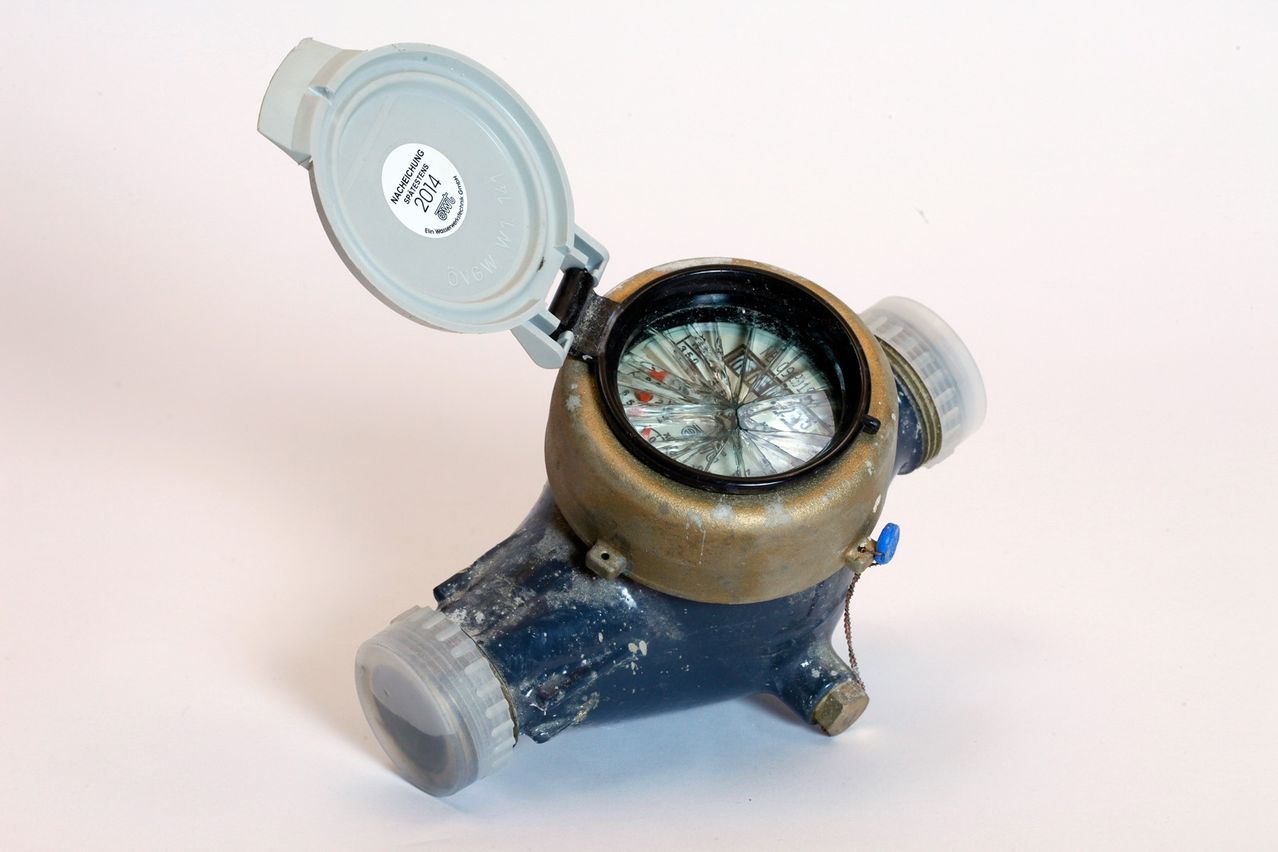 Ein alter, blauer und messingfarbener Wasserzähler mit offenem Deckel, der einen Ziffernblatt mit verschiedenen Zahlen und Markierungen zeigt. Der Deckel hat einen Aufkleber mit dem Text 'NAHEHUNG 2014'.
