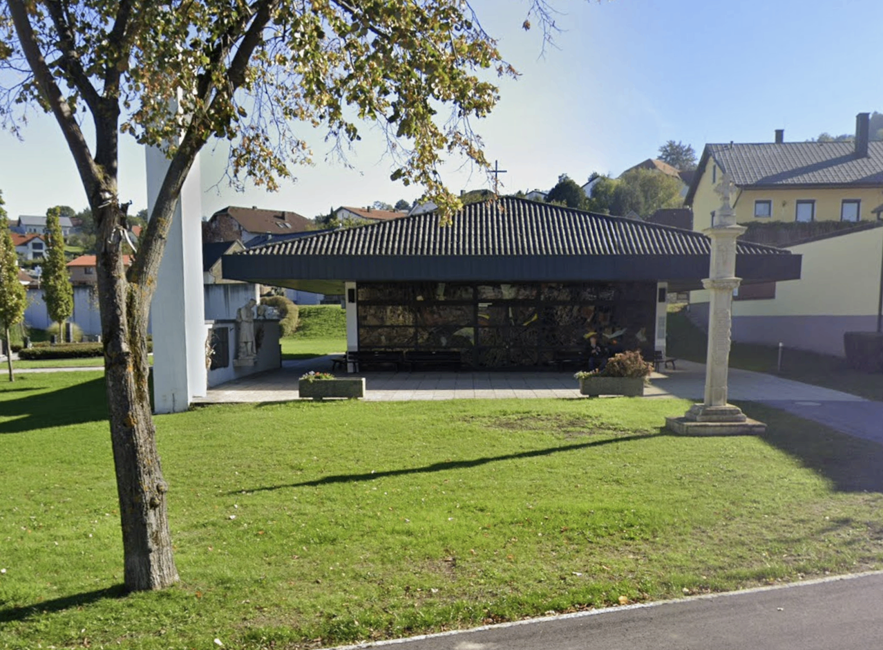 Eine Kapelle mit einem gefliesten Dach und einer mosaikverzierten Wand steht in einer üppigen grünen Landschaft. Sie grenzt an ein weißes Gebäude und wird von einem großen Baum eingerahmt.
