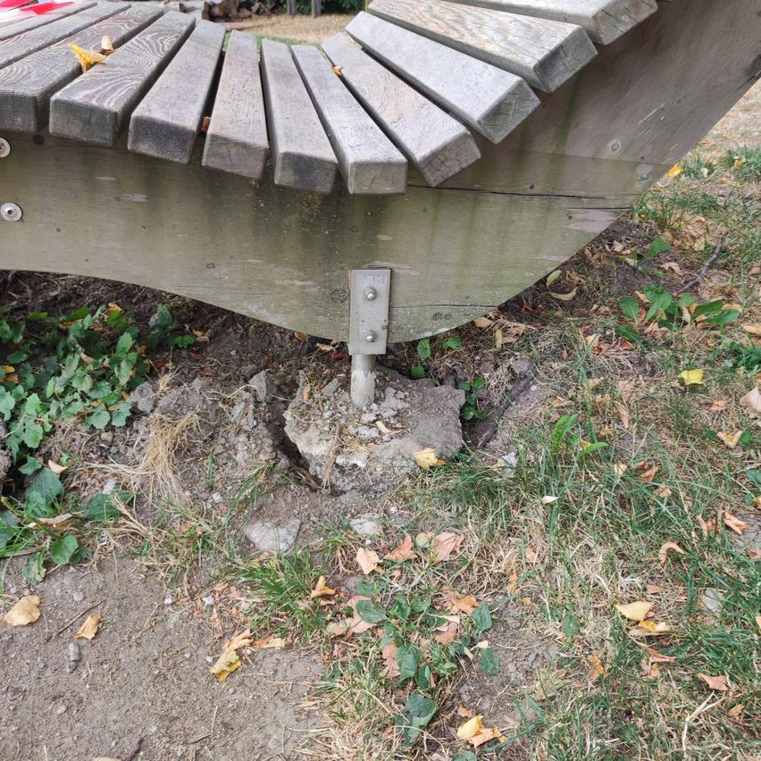 Eine Holzbank mit einem Metallbügel, der an einem Betonsockel befestigt ist, umgeben von Gras und gefallenen Blättern.