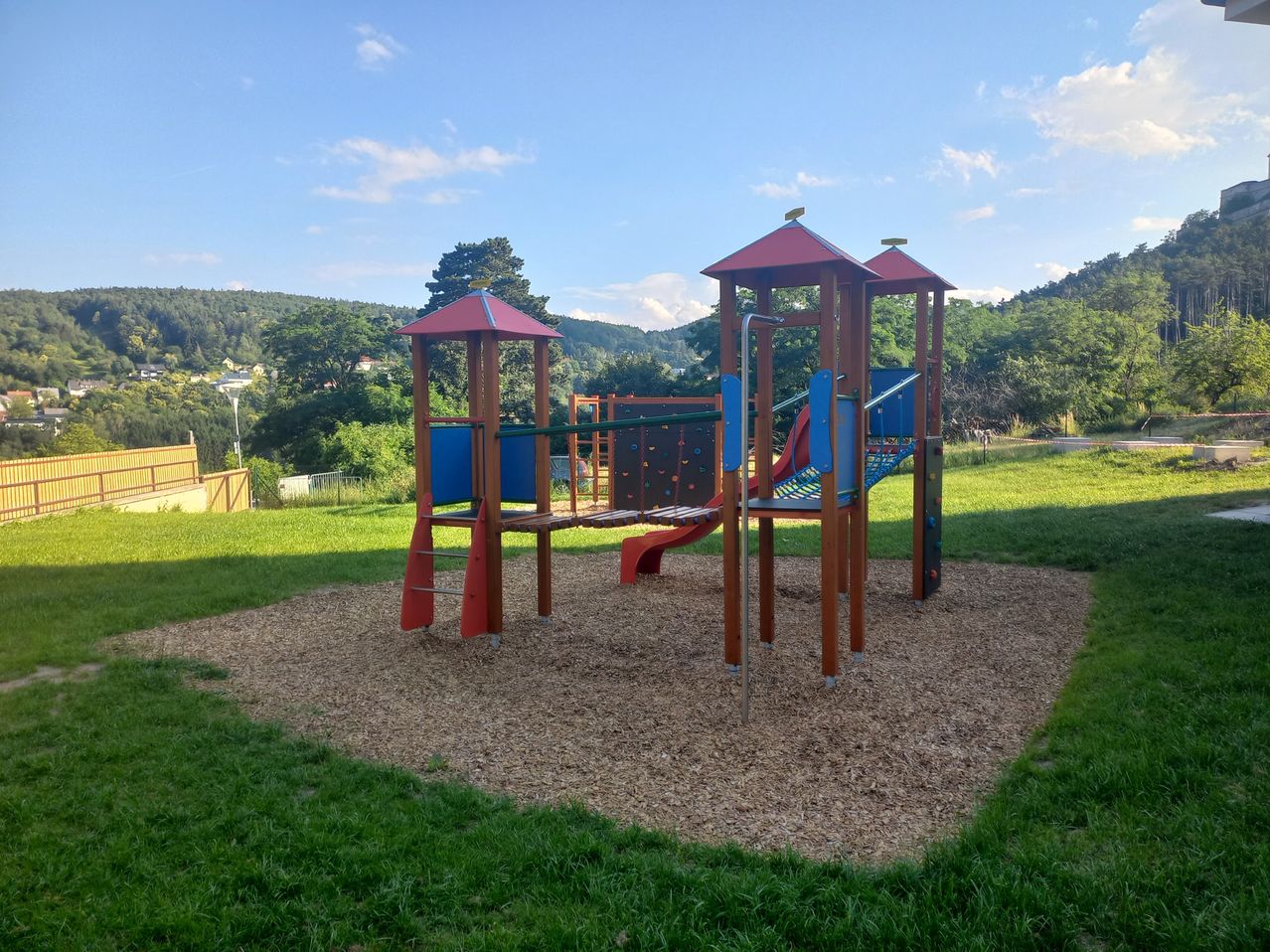 Ein bunter Spielplatz mit einer roten Rutsche und Kletterwand, umgeben von grünem Gras unter einem blauen Himmel mit Wolken.