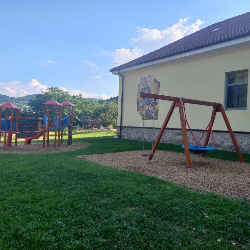 Ein Spielplatz mit Schaukel und Klettergerüst unter blauem Himmel. Hinter ihm steht ein Gebäude mit einer Wandmalerei.