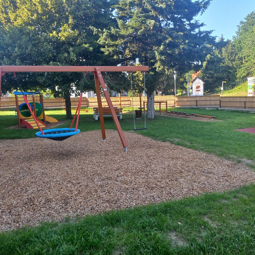 Ein Spielplatz mit Schaukeln, einer Rutsche und einer Holzbank, umgeben von einem Holzzaun unter Bäumen.
