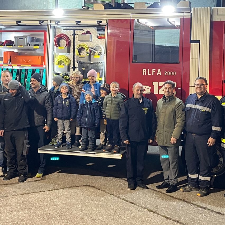 Eine Gruppe von Erwachsenen und Kindern steht vor einem roten Feuerwehrauto. Das Auto ist vor einem Gebäude mit Lichtern in der Nacht geparkt.