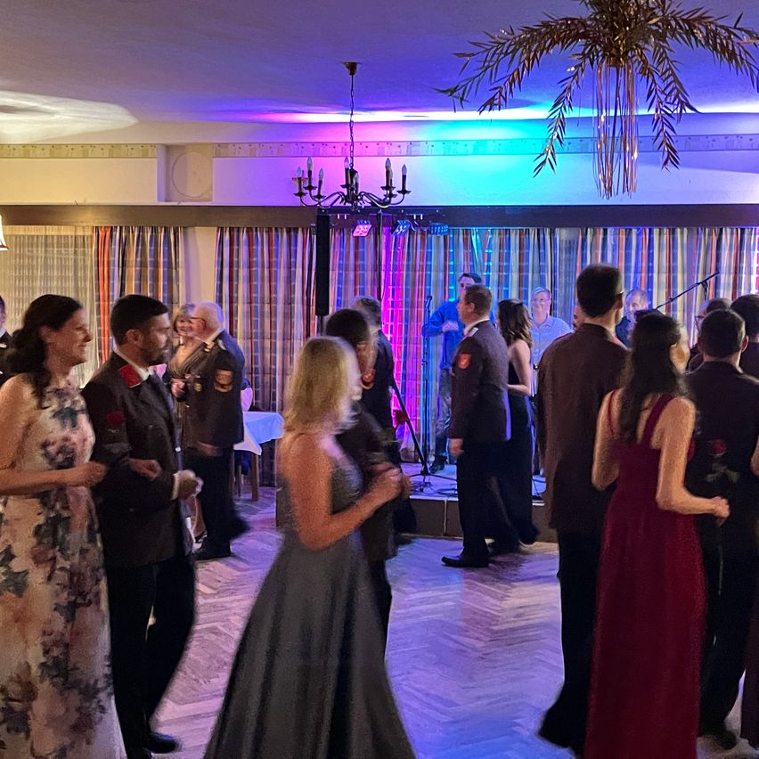 Ein Raum mit Bühne, beleuchtet von rosa und blauen Lichtern. Eine Frau in einem grauen Kleid tanzt, während andere reden oder zusehen. Es gibt Menschen in Uniformen, eine Kronleuchter und Vorhänge.