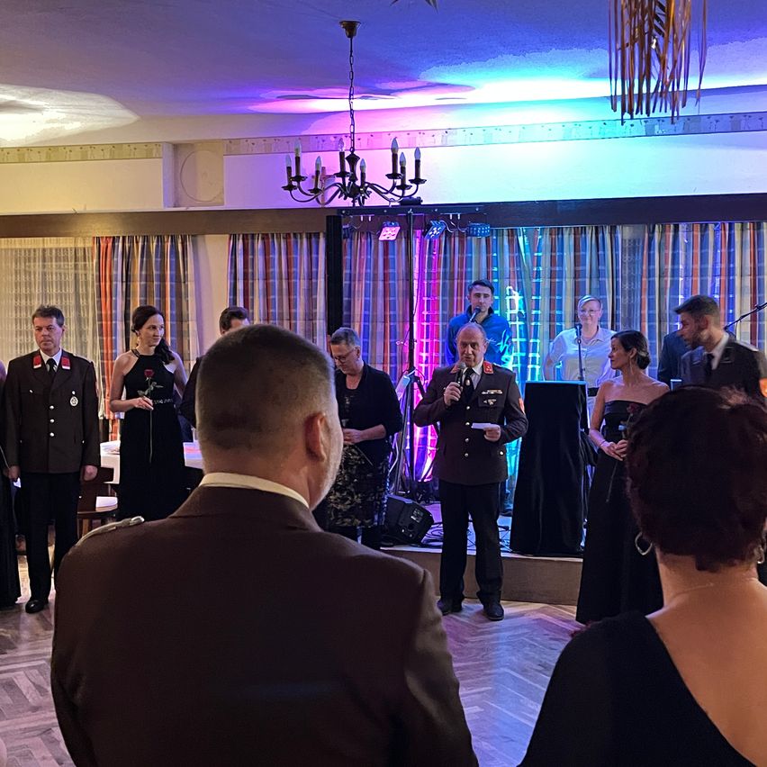 Ein Mann spricht an einem Podium, hält ein Mikrofon in der Hand, während eine Menge uniformierter Offiziere und Gäste steht und zuschaut in einem dekorierten Raum.
