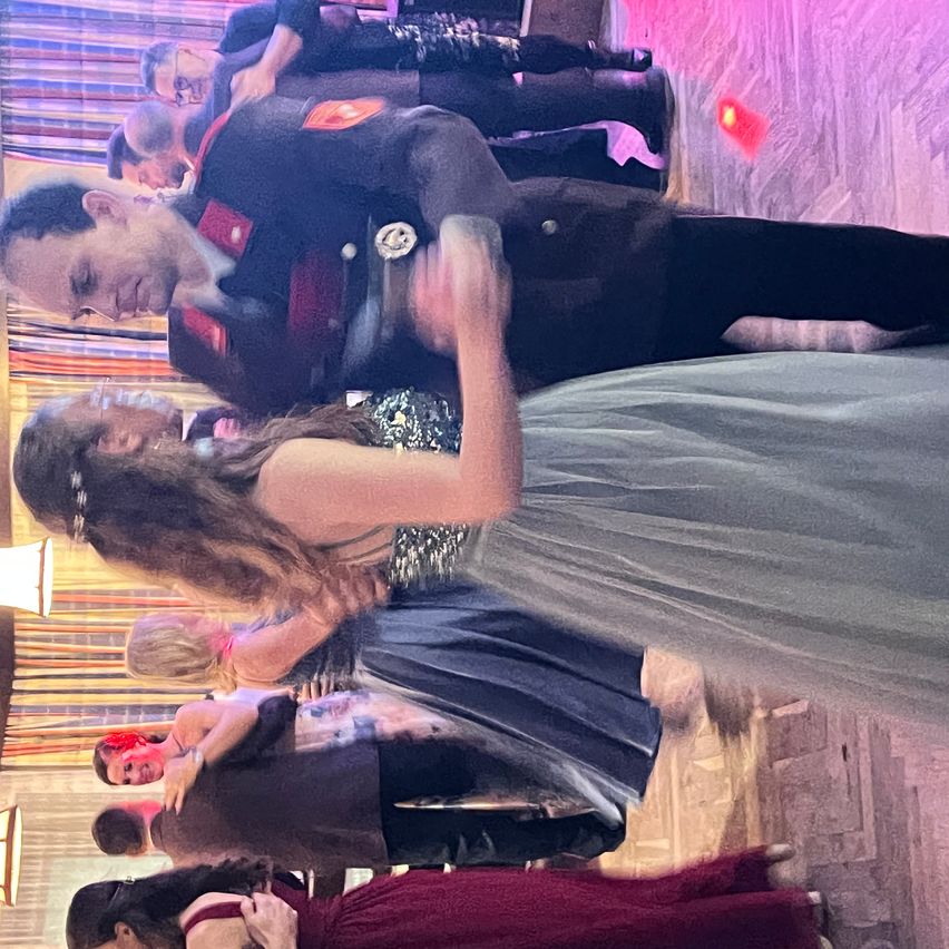 Eine Gruppe von Menschen tanzt in einem Raum. Ein Mann und eine Frau tanzen eng zusammen, mit anderen im Hintergrund. Der Boden ist hölzern und die Wände sind lila.