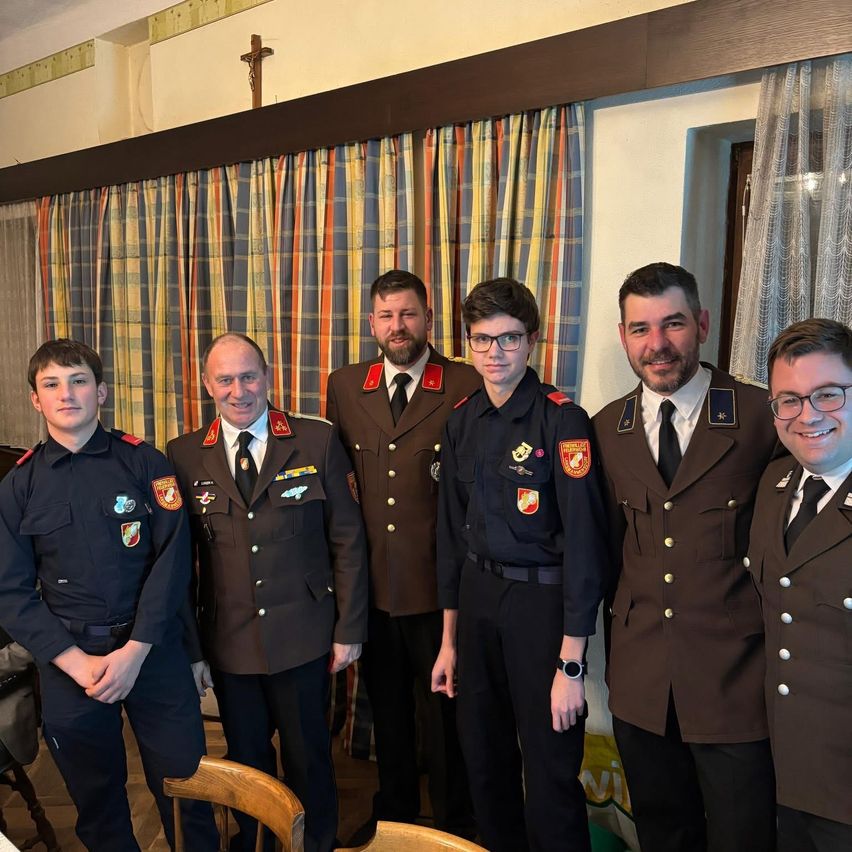 Sechs Männer in Militäruniformen stehen zusammen in einem Raum. Sie lächeln und scheinen für ein Foto zu posieren. Hinter ihnen befinden sich zwei Vorhänge und ein Kreuz.