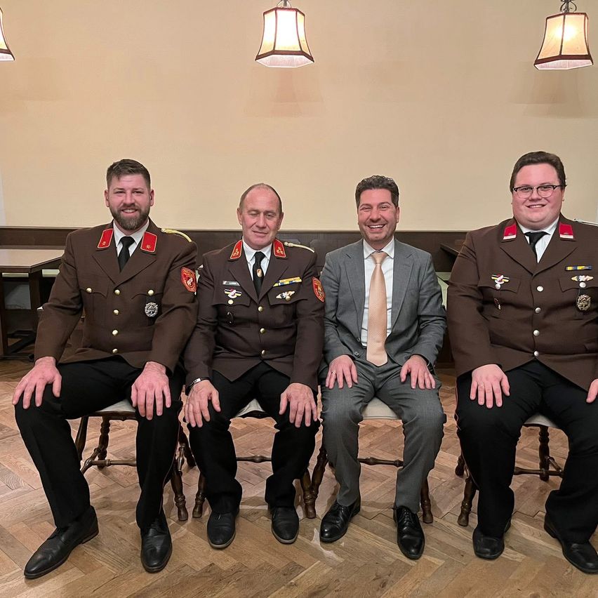 Vier Männer in formeller Kleidung, drei in Militäruniformen und einer in einem Anzug, sitzen zusammen, lächeln, in einem Raum mit Holzboden und hängenden Lampen.