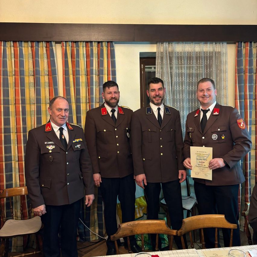 Vier Männer in Militäruniformen stehen lächelnd zusammen, einer hält eine Urkunde. Sie sind in einem Raum mit Holzstühlen und gestreiften Vorhängen.
