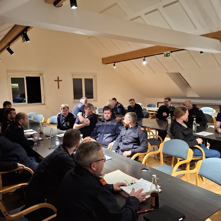 Eine Gruppe uniformierter Männer sitzt um Tische in einem Besprechungsraum herum. Einige schauen sich Papiere an, andere unterhalten sich. Der Raum hat einen Holzboden und weiße Wände.