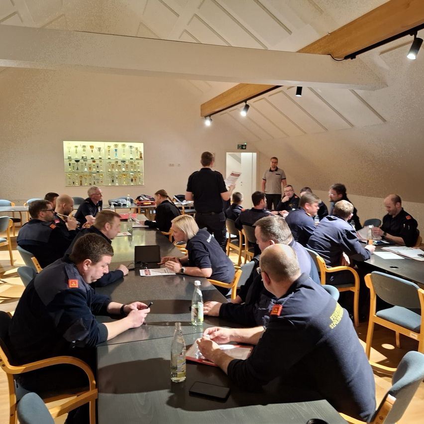 In einem Raum mit einem langen Tisch findet eine Besprechung statt. Mehrere Personen sitzen am Tisch, und eine Person steht in der Nähe des Tisches. Flaschen, Papiere und ein Laptop liegen auf dem Tisch. Eine Lampe ist an der Decke montiert. Eine wandmontierte Anzeige mit Schlüsseln befindet sich links.