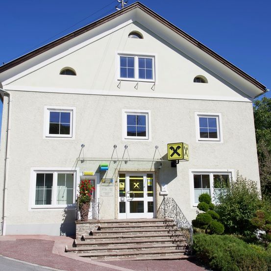 Ein weißes Haus mit einem braunen Dach, mehreren Fenstern und einer Tür mit einem gelben Kreuzsymbol. Das Haus hat eine Treppe, die zum Eingang führt, mit einem Topf und Blumen an der Seite.