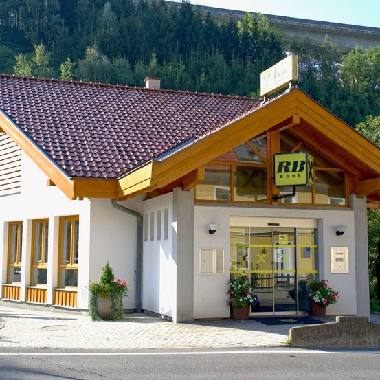 Eine kleine Bankfiliale mit braunem Dach und gelbem Schild, vor einer Straße gelegen, mit einer Brücke im Hintergrund. Der Eingang hat Glastüren und wird von Topfpflanzen flankiert.