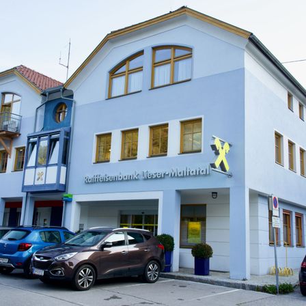 Ein weißes Gebäude mit gelben Akzenten ist die Raffeisenbank Lieser-Maltatal. Autos sind vor dem Gebäude geparkt. Das Gebäude hat Fenster, ein Schild und eine Lampe über dem Eingang.