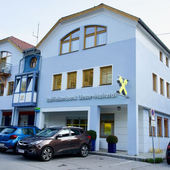 Ein weißes Gebäude mit gelben Akzenten ist die Raffeisenbank Lieser-Maltatal. Autos sind vor dem Gebäude geparkt. Das Gebäude hat Fenster, ein Schild und eine Lampe über dem Eingang.