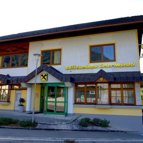 Das Gebäude der Raiffeisenbank Lieser-Maltatal hat ein gelbes und weißes Äußeres mit einem schwarzen Dach. Der Eingang hat grüne Türen und Glasfenster. Ein Schild trägt den Schriftzug 'Raiffeisenbank Lieser-Maltatal'.