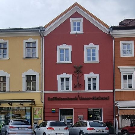 Ein rotes Gebäude mit dem Schild Raiffeisenbank Wasser-Matrei befindet sich zwischen zwei anderen Gebäuden. Vor den Gebäuden sind drei Autos geparkt.