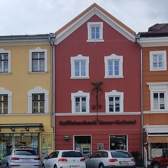 Ein rotes Gebäude mit dem Schild Raiffeisenbank Wasser-Matrei befindet sich zwischen zwei anderen Gebäuden. Vor den Gebäuden sind drei Autos geparkt.