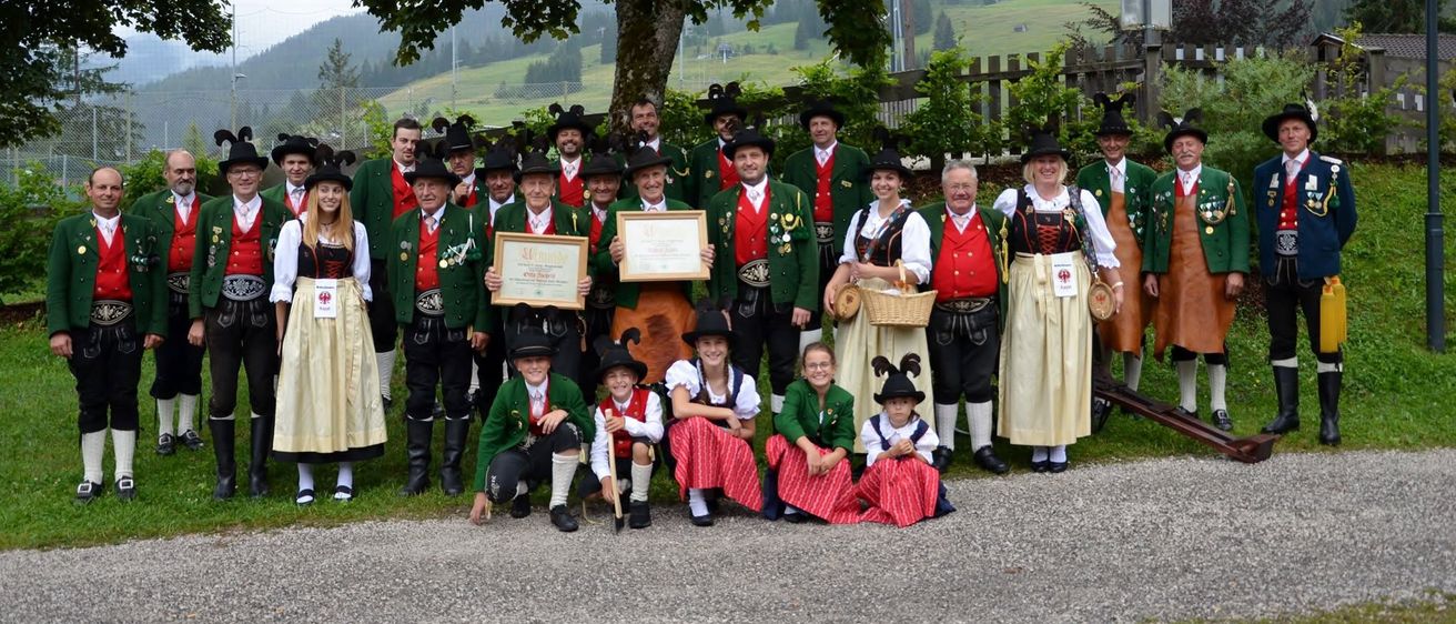 Eine Gruppe von Menschen in traditioneller Kleidung, möglicherweise für ein Fest, posiert für ein Foto unter einem Baum. Einige halten Auszeichnungen, und einige Kinder sind vorne.
