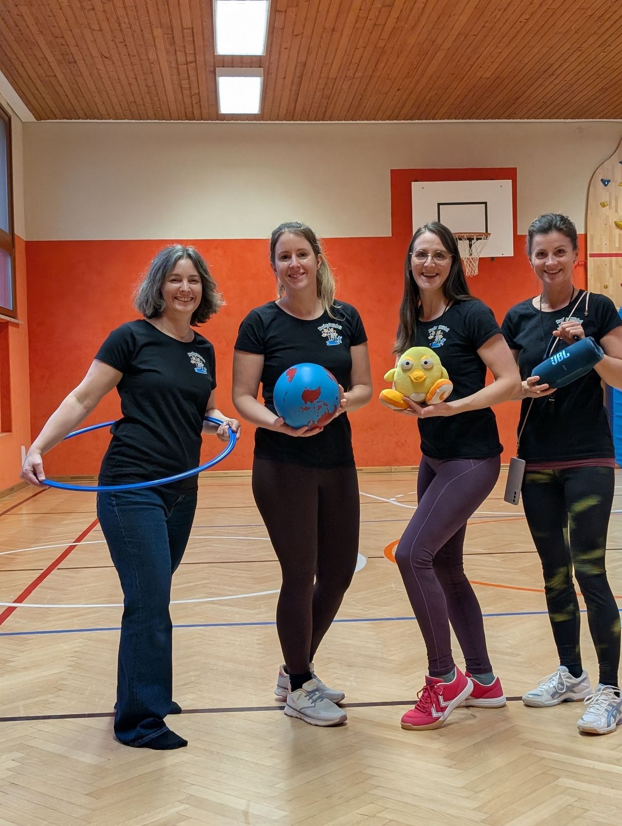 Four women are smiling and posing for a photo in a gym. They are holding a hula hoop, a ball, a stuffed animal, and a water bottle. They are dressed in matching black shirts and leggings. Behind them is a basketball hoop and a climbing wall.
