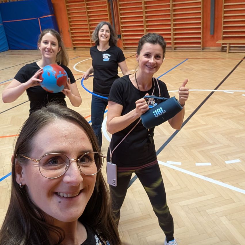 Four women in a gym, three holding exercise equipment, one with a globe, smiling and posing for a photo.