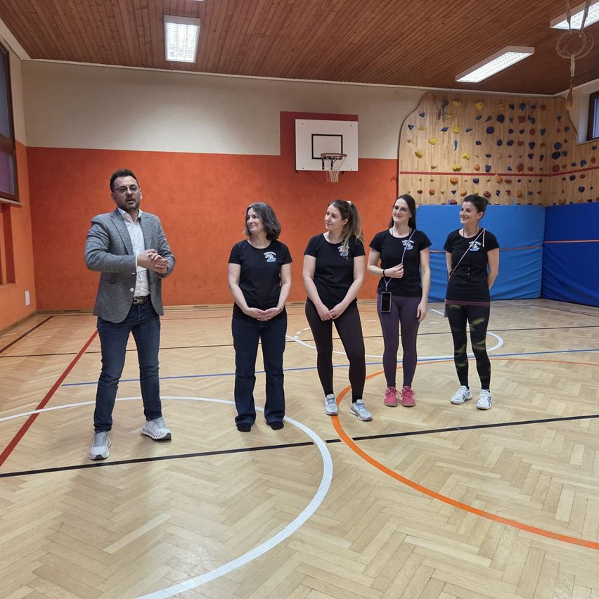 A man and four women stand in a gymnasium, wearing black t-shirts. The man wears glasses and a gray suit. Behind them is a basketball hoop, and a climbing wall is on the wall.