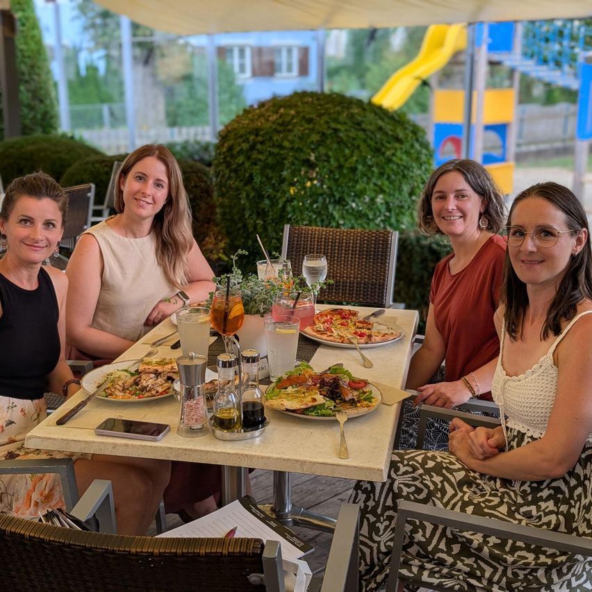 Four women are sitting at an outdoor table, smiling and posing for a photo. The table is set with plates of food, drinks, and utensils. Behind them, there is a hedge and a playground.