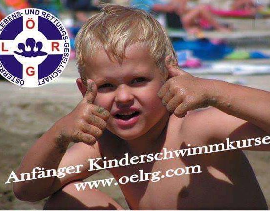 Ein lächelnder junger Junge posiert für ein Foto an einem Sandstrand, trägt ein Schwimmkostüm und ist mit Sand bedeckt. Hinter ihm befindet sich ein Logo mit der Aufschrift 'Lebens-und Rettung-Gesellsschaft'. Im Hintergrund sind verschwommene Bilder von Menschen am Strand zu sehen.