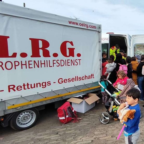 Eine Gruppe von Menschen, darunter Kinder, versammelt sich um einen mit L.R.G. Rettungs-Gesellschaft beschrifteten Lastwagen, einige tragen Rucksäcke. Ein Kind hält einen Plüschtier.