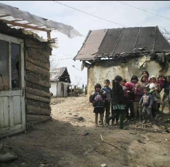 Eine Gruppe von Kindern und Erwachsenen steht vor zwei alten und beschädigten Häusern auf einer unbefestigten Straße. Der Himmel ist bewölkt.