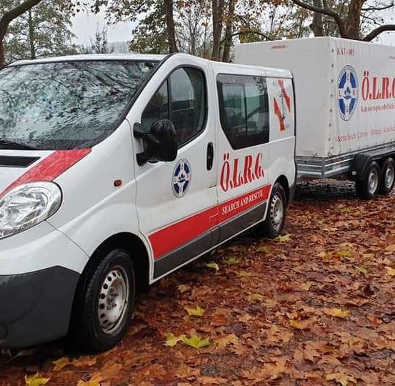 Ein weißer Lieferwagen mit roten Akzenten und der Aufschrift O.L.R.G. steht auf einem laubbedeckten Boden mit angehängtem Anhänger.