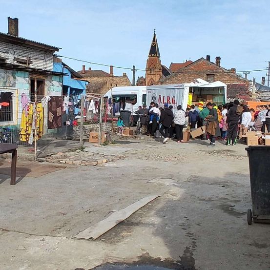 Eine Menschenmenge versammelt sich in einem Viertel, wo ein weißer Lastwagen mit den Buchstaben OLRG geparkt ist. Kisten liegen verstreut auf dem Boden. Menschen erhalten Gegenstände. Im Hintergrund befinden sich Gebäude und ein Turm.