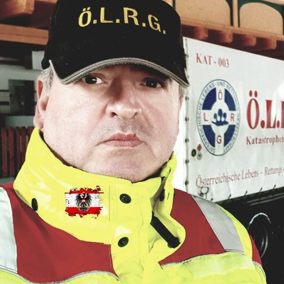 Ein Mann in einer gelben Warnweste, möglicherweise ein Rettungssanitäter, steht vor einem weißen LKW mit Logo und Text. Er trägt eine schwarze Kappe mit gelben Buchstaben und hat eine österreichische Flagge an seiner Jacke befestigt.