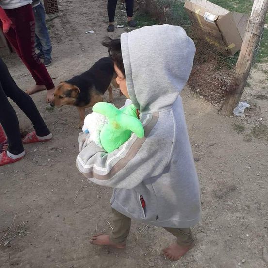 Ein barfuß gehendes Kind mit einem grünen Plüschtier in einer grauen Kapuzenjacke läuft auf einem Erdweg mit Menschen und einem Hund im Hintergrund.