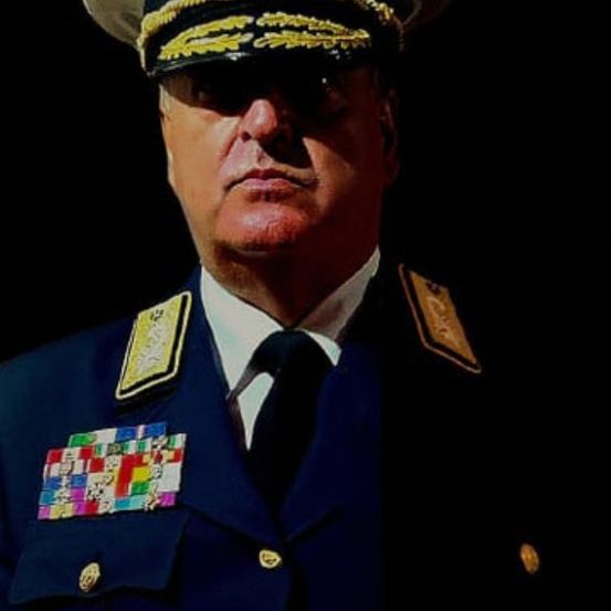 Ein Militäroffizier steht mit strengem Gesichtsausdruck, trägt eine blaue Uniform, eine schwarze Krawatte und einen Hut mit goldenen Verzierungen. Mehrere Medaillen schmücken seine Brust.