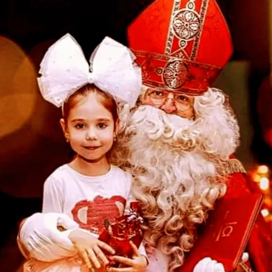 Ein kleines Mädchen mit einer weißen Schleife im Haar hält ein Geschenk und lächelt, während es neben einem Mann steht, der als Weihnachtsmann verkleidet ist.