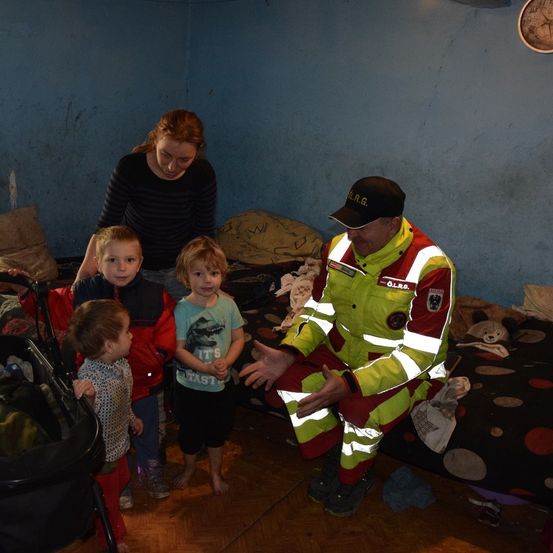 Ein Feuerwehrmann spricht mit einer Frau und vier Kindern in einem Zimmer. Die Frau hält ein Kind. Die Kinder tragen Jacken. Ein Kinderwagen steht in der Nähe.