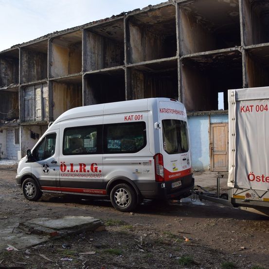 Ein weißer Van mit roten Akzenten und den Buchstaben O.L.R.G. ist vor einem großen, baufälligen Gebäude geparkt. Der Van hat KAT 001 auf der Rückseite.