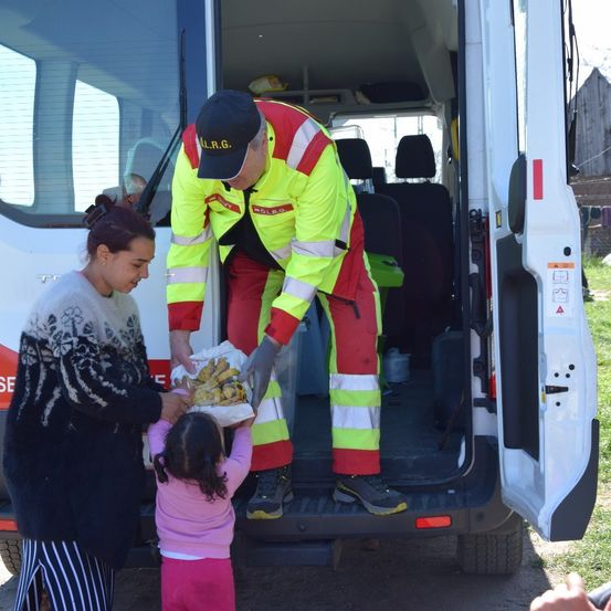 Eine Person in einer reflektierenden Weste reicht einer Frau und einem Kind vor einem Lieferwagen eine Tüte mit Essen.