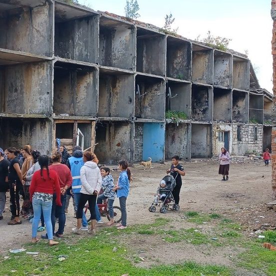 Eine Gruppe von Menschen steht vor einem verlassenen, beschädigten Gebäude mit vielen leeren Räumen und einem Hund in der Nähe.
