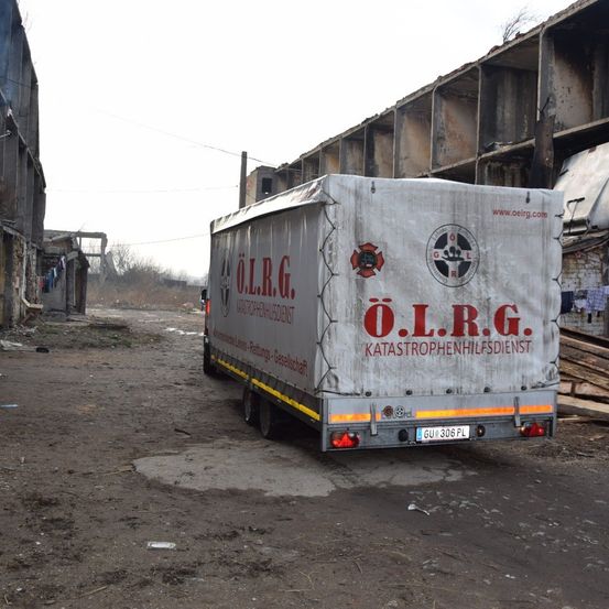 Ein weißer Anhänger mit der Aufschrift O.L.R.G. Katastrophenhilfedienst ist auf einer unbefestigten Straße vor einem beschädigten Gebäude geparkt.