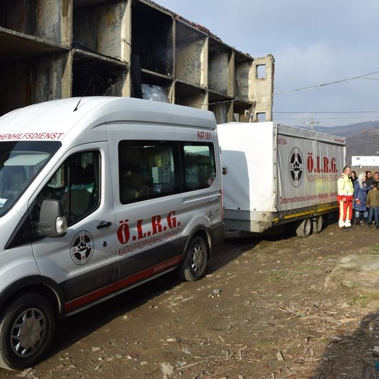 Ein weißer Van mit rotem Text steht neben einem Anhänger mit Logo und einer Gruppe von Personen in Sicherheitsanzügen.