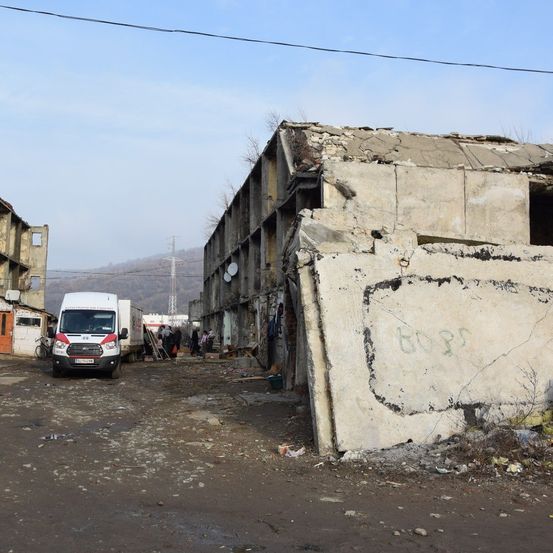 Ein beschädigtes Gebäude steht neben einer Straße mit einem geparkten Krankenwagen, Menschen versammeln sich in der Nähe und eine große Wand hat Graffiti.