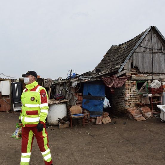 Ein Mann in einer Schutzkleidung und Gesichtsmaske geht an einem beschädigten Gebäude mit einem beschädigten Dach und verstreuten Gegenständen vorbei.