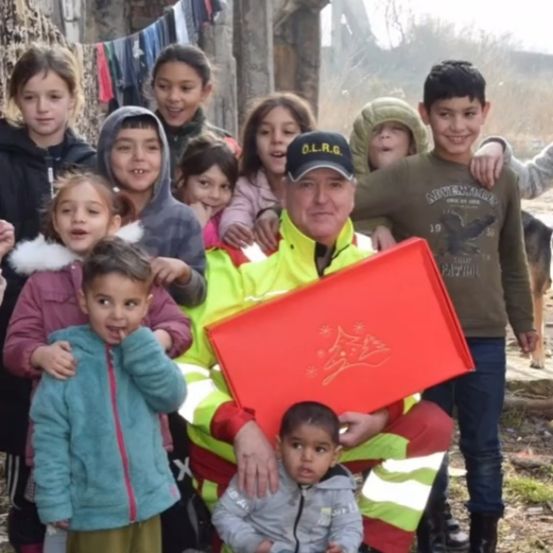 Ein Mann in reflektierender Kleidung hält eine rote Box, umgeben von Kindern in verschiedenen Jacken, alle lächeln und posieren für ein Foto.