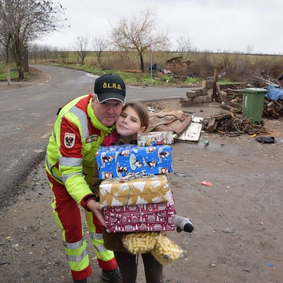 Ein Mann in einer Warnweste hält ein junges Mädchen mit Weihnachtsgeschenken in einem beschädigten Gebiet.